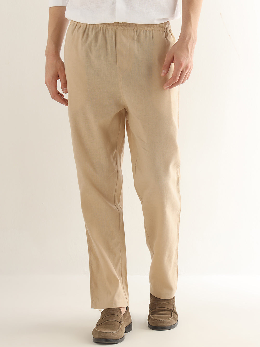 A person wearing beige straight-leg trousers with an elastic waistband, paired with brown suede loafers and a white shirt, shown from the waist down against a plain light background.