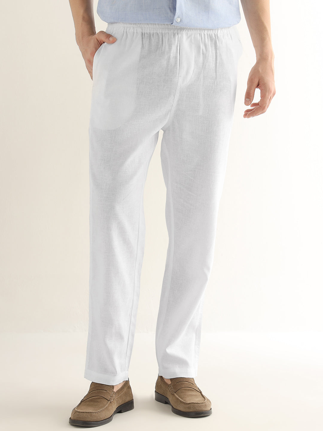 White linen trousers with an elastic waistband, modeled with a light blue shirt and brown suede loafers against a warm off-white background.